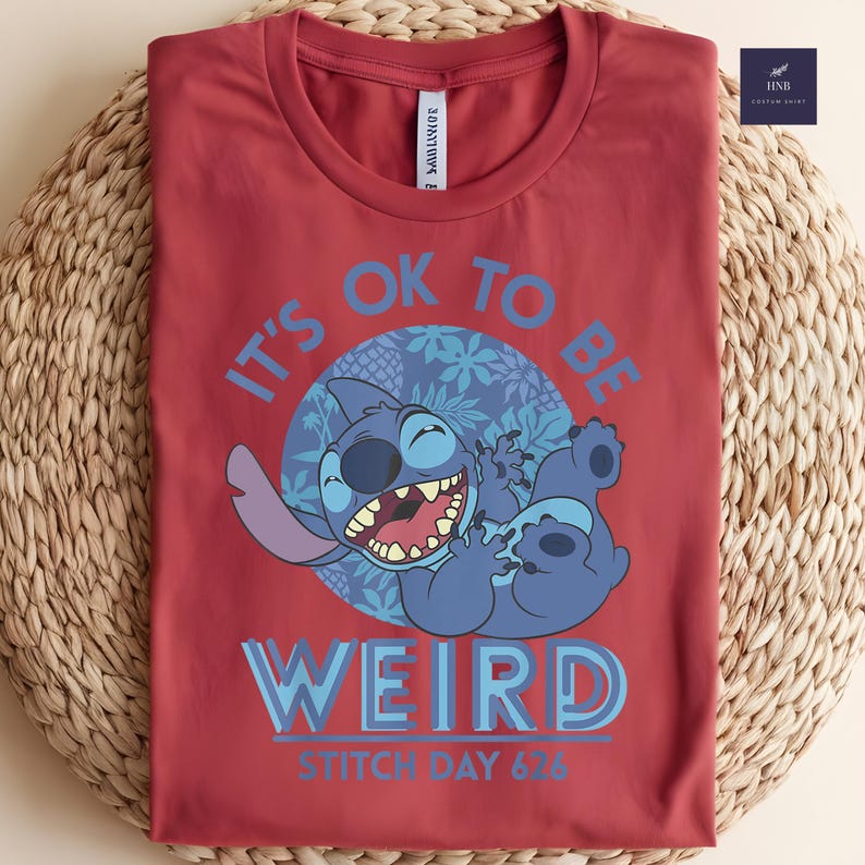 Puede incluir: Camiseta roja con un personaje de dibujos animados azul y el texto "IT'S OK TO BE WEIRD" y "STITCH DAY 626". El personaje est&aacute; sonriendo y tumbado. La camiseta est&aacute; doblada sobre una superficie tejida.