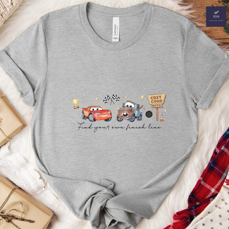 May include: A gray t-shirt featuring a graphic of two cartoon cars, a red race car and a brown tow truck, with the text "Find your own finish line." The design includes a trophy, checkered flags, and a motel sign.