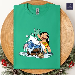 Pode incluir: Camiseta verde com uma ilustra&ccedil;&atilde;o de desenho animado de Lilo lavando Stitch em uma banheira de madeira. Lilo est&aacute; usando um mai&ocirc; listrado amarelo e laranja. Stitch &eacute; azul e coberto de espuma branca. A camiseta tem gola redonda.