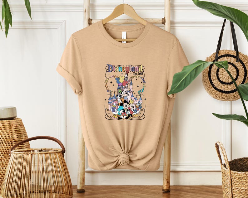 Może przedstawiać: Beżowy t-shirt z kr&oacute;tkim rękawem z nadrukiem graficznym przedstawiającym zamek Disneyland, Myszkę Miki i inne postacie z kresk&oacute;wek. Napis "Disneyland Est. 1955" znajduje się nad zamkiem i jest wykonany kolorową, stylizowaną czcionką z gwiazdkami.