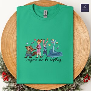 K&ouml;nnte beinhalten: Ein gr&uuml;nes T-Shirt mit einem Cartoon-Design eines Bibers, Fuchses, Kaninchens und einer Schlange mit dem Text "Anyone can be anything". Das Shirt wird auf einem Holztablett mit dekorativen Elementen pr&auml;sentiert.