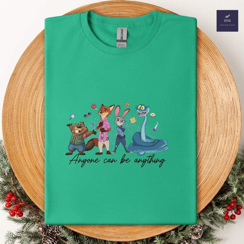 K&ouml;nnte beinhalten: Ein gr&uuml;nes T-Shirt mit einem Cartoon-Design eines Bibers, Fuchses, Kaninchens und einer Schlange mit dem Text "Anyone can be anything". Das Shirt wird auf einem Holztablett mit dekorativen Elementen pr&auml;sentiert.