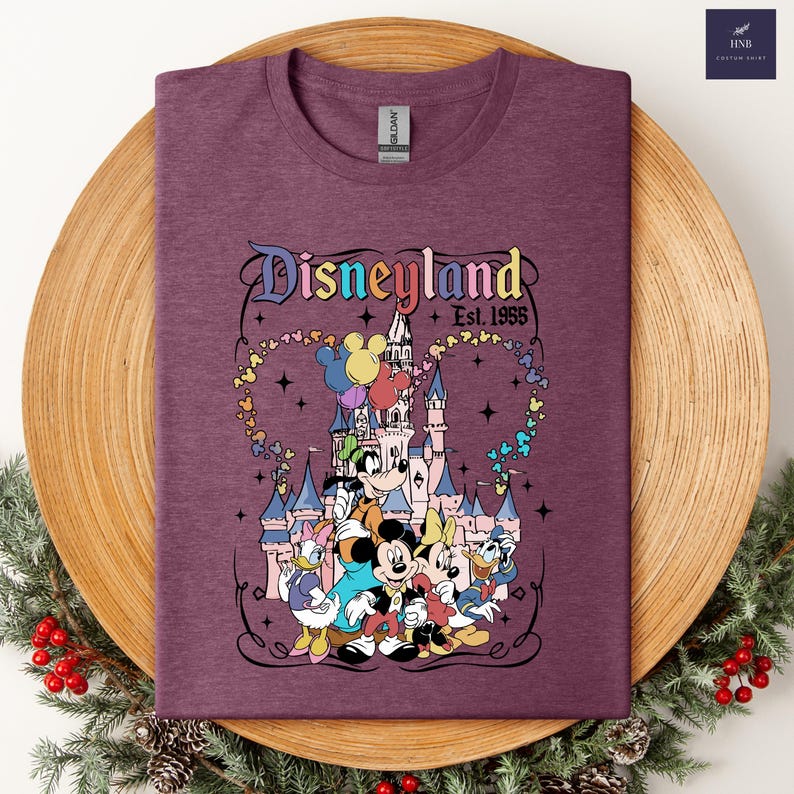 Może przedstawiać: Melirowany bordowy t-shirt z grafiką zamku Disneyland oraz postaciami Myszki Miki, Myszki Minnie, Goofy'ego, Kaczora Donalda i Daisy. Napis głosi Disneyland Est. 1955, otoczony kolorowym szlakiem balon&oacute;w w kształcie głowy Myszki Miki tworzących serce.