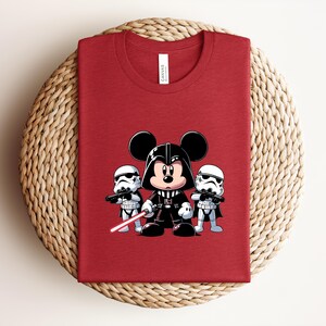 Pu&ograve; includere: T-shirt rosso con un'illustrazione a cartoni animati di Topolino nei panni di Darth Vader, affiancato da due Stormtrooper. Il disegno include una spada laser rossa. La maglietta &egrave; piegata su una superficie intrecciata.