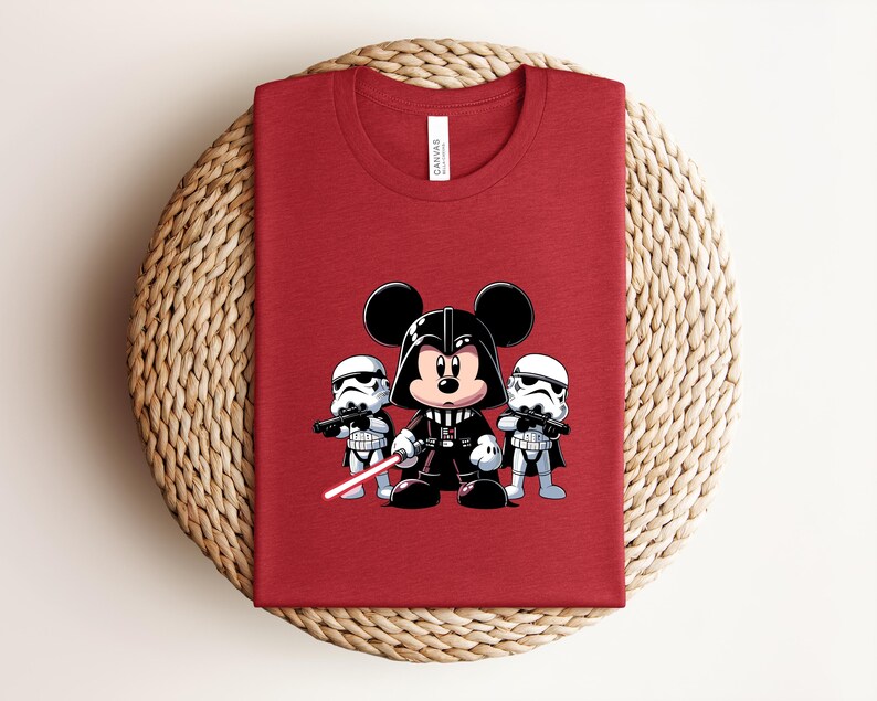 Pu&ograve; includere: T-shirt rosso con un'illustrazione a cartoni animati di Topolino nei panni di Darth Vader, affiancato da due Stormtrooper. Il disegno include una spada laser rossa. La maglietta &egrave; piegata su una superficie intrecciata.