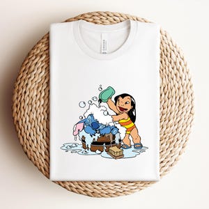 Pode incluir: Camiseta branca com uma ilustra&ccedil;&atilde;o de desenho animado de Lilo e Stitch. Lilo est&aacute; despejando &aacute;gua em Stitch, que est&aacute; em uma banheira de madeira cheia de bolhas. A camiseta est&aacute; dobrada em uma superf&iacute;cie circular tecida.