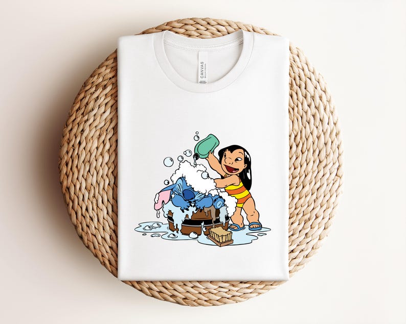 Pode incluir: Camiseta branca com uma ilustra&ccedil;&atilde;o de desenho animado de Lilo e Stitch. Lilo est&aacute; despejando &aacute;gua em Stitch, que est&aacute; em uma banheira de madeira cheia de bolhas. A camiseta est&aacute; dobrada em uma superf&iacute;cie circular tecida.