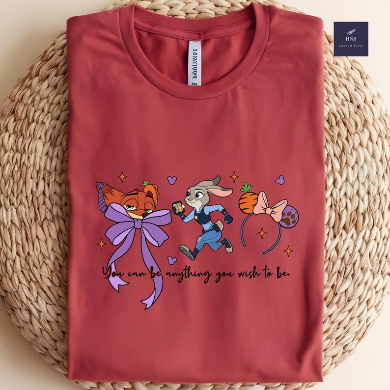 May include: A maroon t-shirt featuring a cartoon design with a fox, a rabbit, and a carrot-shaped headband. The text "You can be anything you wish to be" is printed below the characters. The shirt has a round neckline.