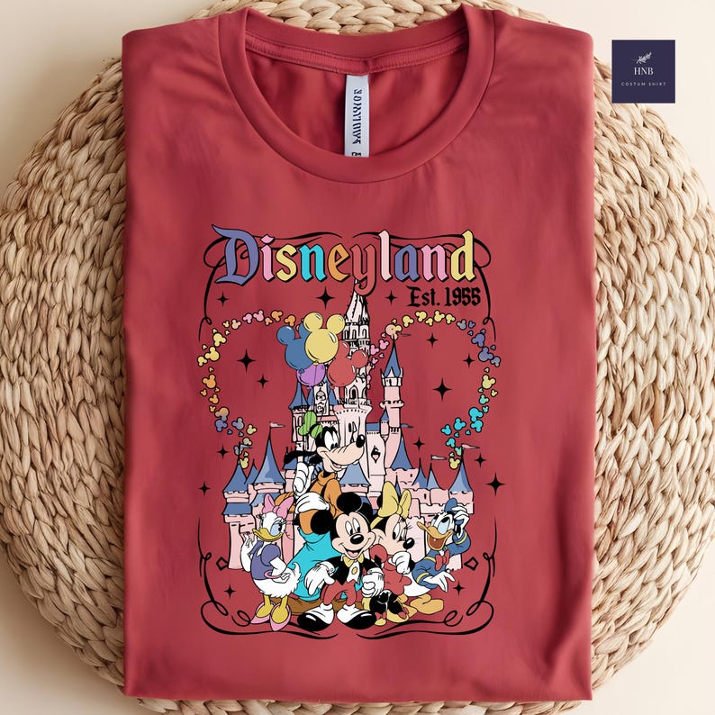 Może przedstawiać: Ceglastoczerwony t-shirt z okrągłym dekoltem i grafiką Disneylandu z napisem Disneyland Est. 1955. Wz&oacute;r przedstawia Myszkę Miki, Myszkę Minnie, Kaczora Donalda, Daisy i Goofy'ego na tle zamku z kolorowymi balonami w kształcie serca.