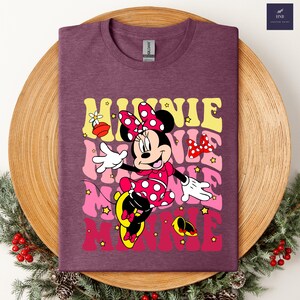 May include: A maroon t-shirt featuring a graphic of Minnie Mouse in a red polka-dot dress and bow. The word "MINNIE" is layered in yellow, pink, and red, with star accents. The shirt is displayed on a wooden tray with holiday greenery.
