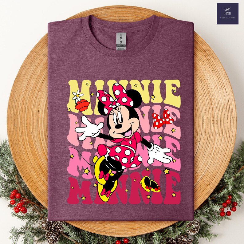 May include: A maroon t-shirt featuring a graphic of Minnie Mouse in a red polka-dot dress and bow. The word "MINNIE" is layered in yellow, pink, and red, with star accents. The shirt is displayed on a wooden tray with holiday greenery.