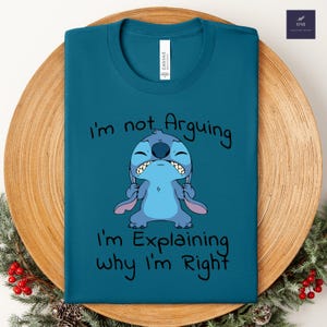 May include: Teal t-shirt featuring a cartoon character with the text "I'm not Arguing, I'm Explaining why I'm Right." The character is blue with large ears and a grumpy expression. The shirt is displayed on a wooden plate.