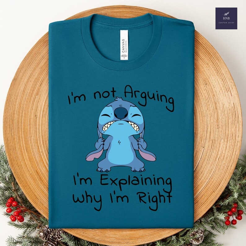 May include: Teal t-shirt featuring a cartoon character with the text "I'm not Arguing, I'm Explaining why I'm Right." The character is blue with large ears and a grumpy expression. The shirt is displayed on a wooden plate.