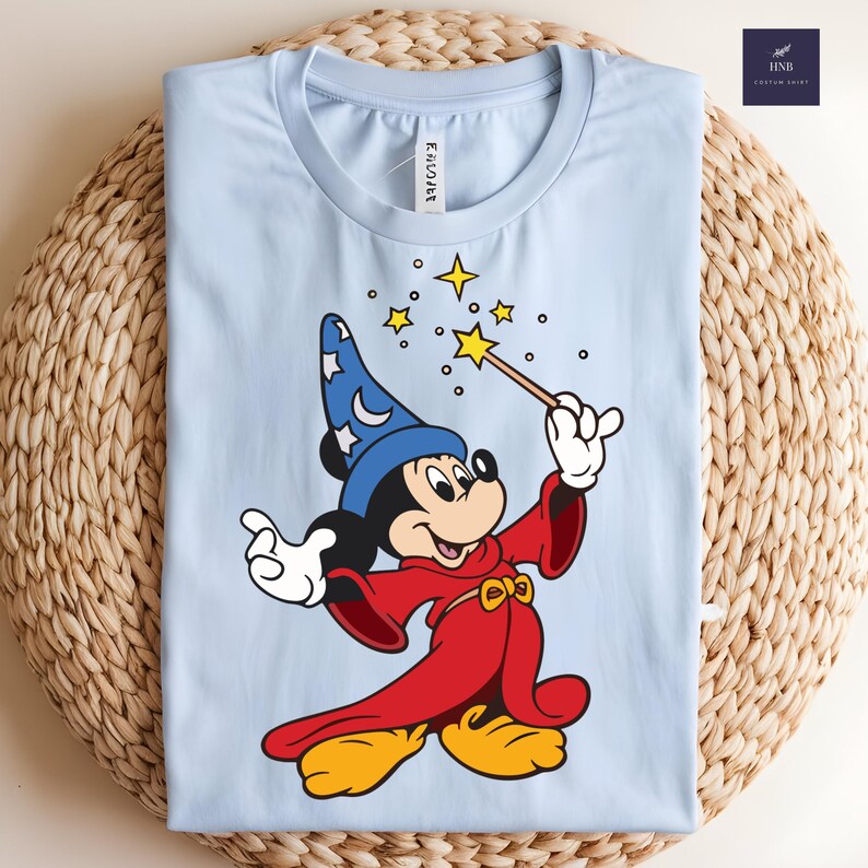 Puede incluir: Camiseta azul claro con Mickey Mouse de dibujos animados vestido de mago. Mickey lleva un sombrero azul con estrellas blancas, una t&uacute;nica roja y zapatos amarillos. Sostiene una varita y conjura estrellas. La camiseta est&aacute; doblada sobre una superficie tejida.