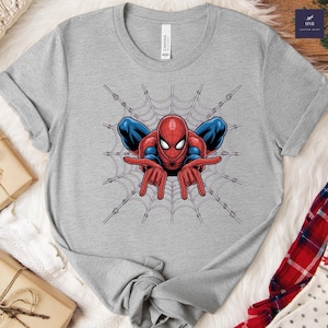 May include: A gray t-shirt featuring a graphic of Spider-Man in a web. The superhero is depicted in his classic red and blue suit, with his hands outstretched. The shirt is made of a soft material and has a crew neck.