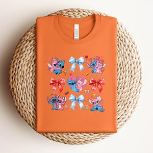 May include: Orange t-shirt featuring a colorful graphic of cartoon characters, bows, and hearts. The design includes blue and pink characters in various poses, with red and blue bows. The shirt is folded on a woven, circular surface.