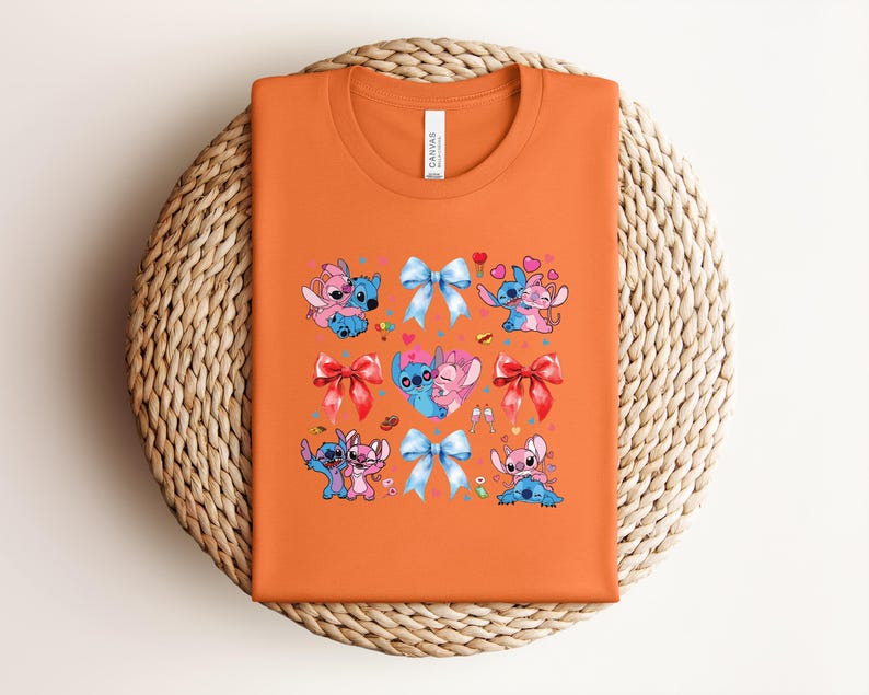 May include: Orange t-shirt featuring a colorful graphic of cartoon characters, bows, and hearts. The design includes blue and pink characters in various poses, with red and blue bows. The shirt is folded on a woven, circular surface.