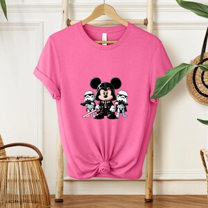 Pu&ograve; includere: Una maglietta rosa acceso con un disegno a cartoni animati di Topolino nei panni di Darth Vader, affiancato da due Stormtrooper. Il design &egrave; in bianco, nero e grigio. La maglietta &egrave; appesa a un appendiabiti di legno.