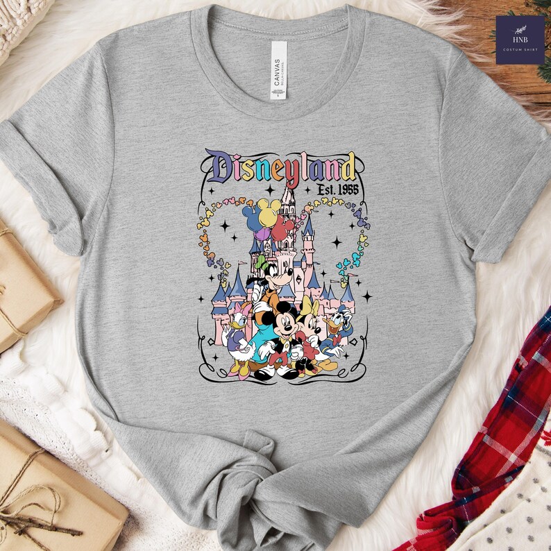 Może przedstawiać: Szary melanżowy t-shirt z kr&oacute;tkim rękawem z nadrukiem graficznym przedstawiającym zamek Disneyland oraz klasyczne postacie z kresk&oacute;wek, w tym Myszkę Miki, Minnie, Kaczora Donalda, Daisy i Goofy'ego, z napisem Disneyland Est. 1955 i kolorowymi sercami.