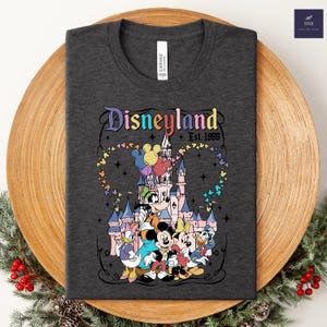 Może przedstawiać: Ciemnoszary t-shirt z kolorową grafiką zamku Disneyland i klasycznymi postaciami z kresk&oacute;wek. Napis brzmi Disneyland Est. 1955, otoczony śladem w kształcie serca złożonym z małych, wielokolorowych ikon uszu Myszki Miki.