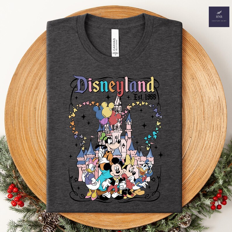 Może przedstawiać: Ciemnoszary t-shirt z kolorową grafiką zamku Disneyland i klasycznymi postaciami z kresk&oacute;wek. Napis brzmi Disneyland Est. 1955, otoczony śladem w kształcie serca złożonym z małych, wielokolorowych ikon uszu Myszki Miki.
