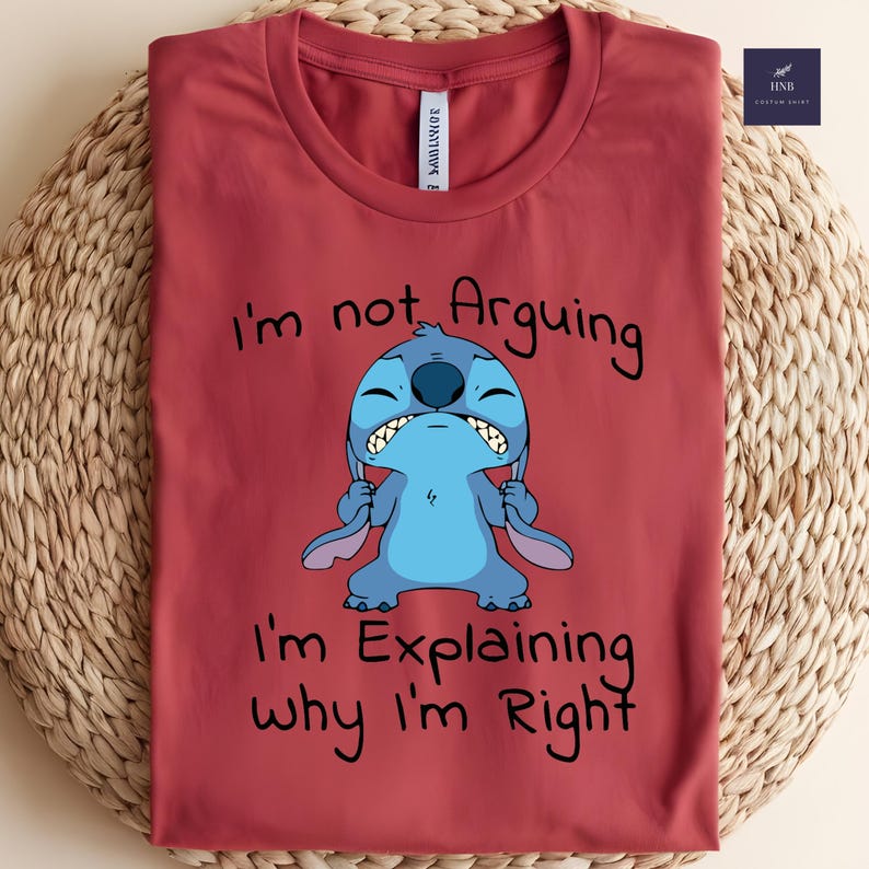 May include: A maroon t-shirt featuring a blue cartoon character with the text "I'm not Arguing, I'm Explaining why I'm Right." The character is depicted with a grumpy expression. The shirt is folded on a woven surface.