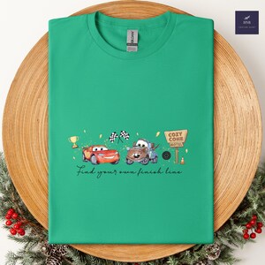 May include: A green t-shirt featuring a cartoon design of two cars, a trophy, and checkered flags. The text "Find your own finish line" is printed below the cars. The shirt is displayed on a wooden plate.