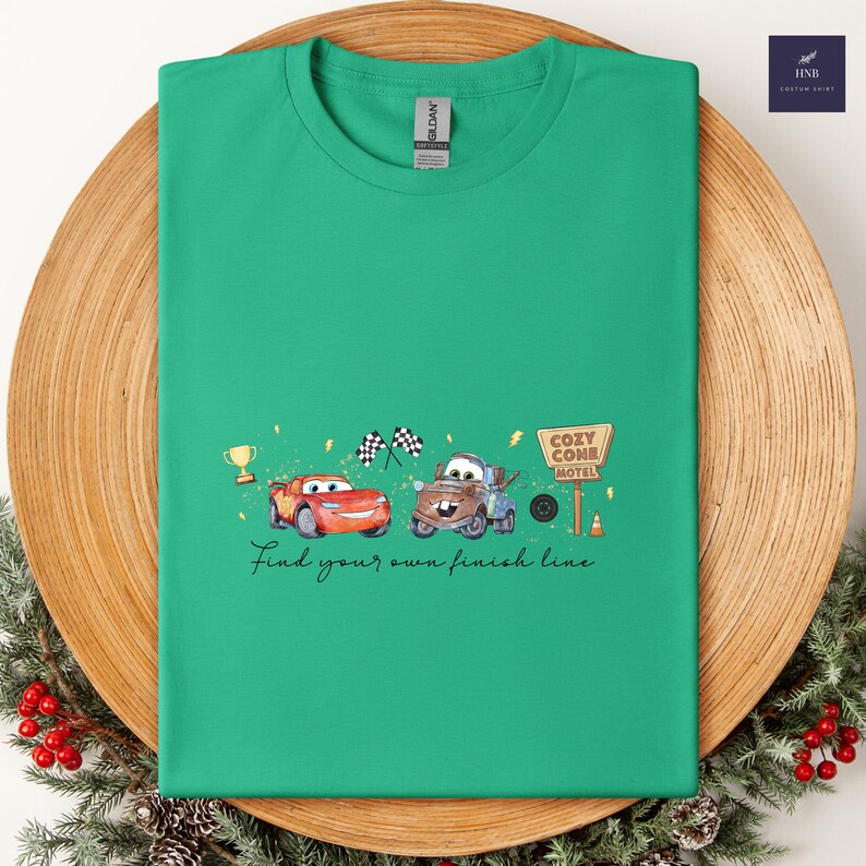 May include: A green t-shirt featuring a cartoon design of two cars, a trophy, and checkered flags. The text "Find your own finish line" is printed below the cars. The shirt is displayed on a wooden plate.