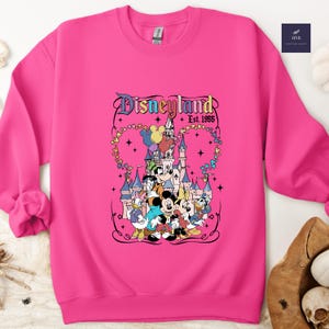 Może przedstawiać: Jaskrawor&oacute;żowa bluza z okrągłym dekoltem i nadrukiem graficznym Disneylandu z napisem 'Disneyland Est. 1955'. Wz&oacute;r przedstawia zamek, Myszkę Miki, Myszkę Minnie, Goofy'ego, Kaczora Donalda i Daisy w otoczeniu kolorowych balon&oacute;w.
