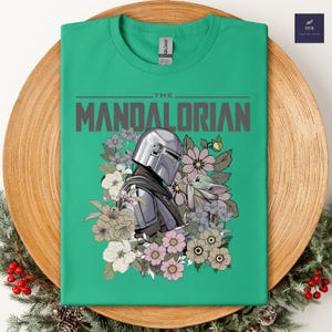 Puede incluir: Camiseta verde con un dise&ntilde;o de The Mandalorian y Grogu rodeados de flores. El dise&ntilde;o incluye el texto "THE MANDALORIAN" en gris. La camiseta se muestra en un plato de madera con vegetaci&oacute;n festiva.