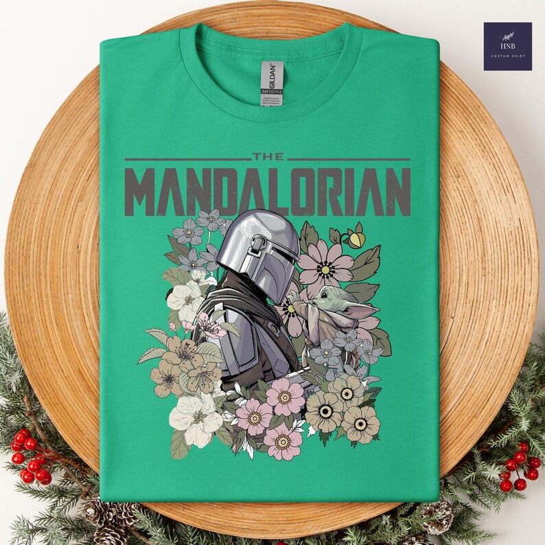 Puede incluir: Camiseta verde con un dise&ntilde;o de The Mandalorian y Grogu rodeados de flores. El dise&ntilde;o incluye el texto "THE MANDALORIAN" en gris. La camiseta se muestra en un plato de madera con vegetaci&oacute;n festiva.