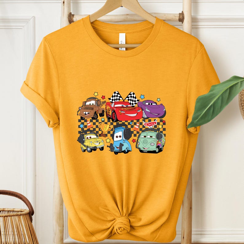 May include: Mustard yellow t-shirt featuring a graphic of cartoon cars. The design includes characters like Mater, Lightning McQueen, and others, with checkered flags and a trophy. The shirt is hanging on a wooden hanger.
