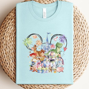 May include: Light blue t-shirt featuring a colorful graphic of Disney characters, including Woody, Buzz Lightyear, and others, arranged within a Mickey Mouse ear outline. The shirt is made of a soft material and is perfect for fans of the animated film.