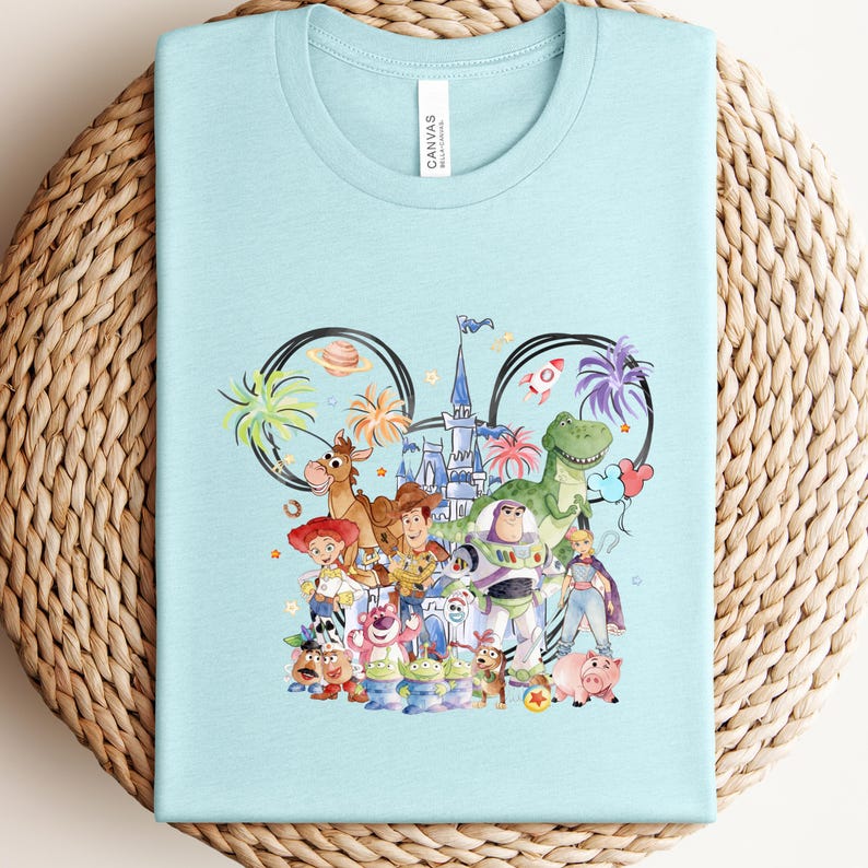 May include: Light blue t-shirt featuring a colorful graphic of Disney characters, including Woody, Buzz Lightyear, and others, arranged within a Mickey Mouse ear outline. The shirt is made of a soft material and is perfect for fans of the animated film.