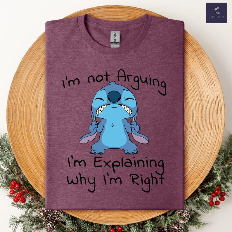 May include: A maroon t-shirt featuring a cartoon character with the text "I'm not Arguing, I'm Explaining why I'm Right." The character is blue with large ears and a grumpy expression. The shirt is displayed on a wooden plate.
