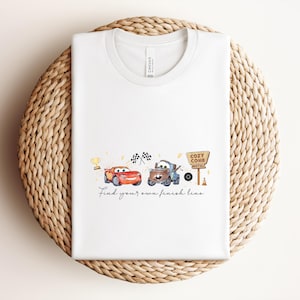 May include: White t-shirt featuring a watercolor design of two cartoon cars, a trophy, and checkered flags. The text "Find your own finish line" is below the cars. The shirt is folded on a woven, circular surface.