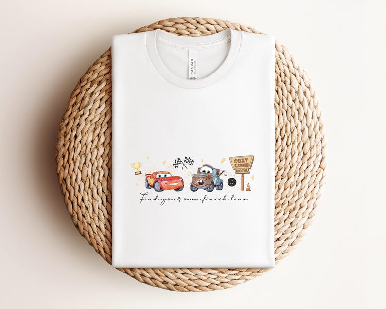 May include: White t-shirt featuring a watercolor design of two cartoon cars, a trophy, and checkered flags. The text "Find your own finish line" is below the cars. The shirt is folded on a woven, circular surface.