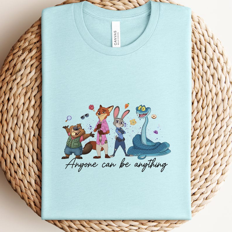 K&ouml;nnte beinhalten: Hellblaues T-Shirt mit einer Cartoon-Illustration von Tierfiguren, darunter ein Biber, ein Fuchs, ein Kaninchen und eine Schlange. Der Text "Anyone can be anything" ist in einer Schriftart unter den Figuren aufgedruckt.