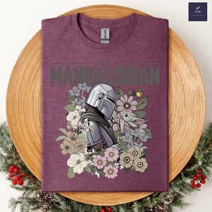 Puede incluir: Una camiseta granate con un gr&aacute;fico del Mandaloriano y Grogu rodeados de flores. La palabra "MANDALORIAN" est&aacute; impresa en la parte superior en gris. La camiseta tiene cuello redondo y una etiqueta Gildan.