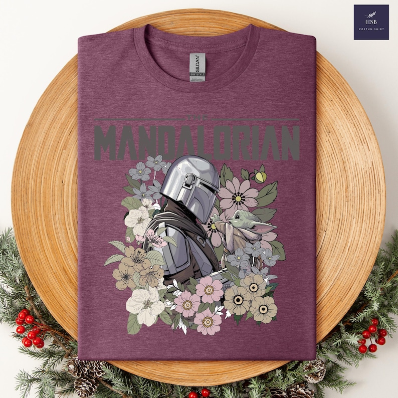 Puede incluir: Una camiseta granate con un gr&aacute;fico del Mandaloriano y Grogu rodeados de flores. La palabra "MANDALORIAN" est&aacute; impresa en la parte superior en gris. La camiseta tiene cuello redondo y una etiqueta Gildan.