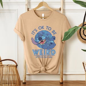 Puede incluir: Camiseta color beige con un dise&ntilde;o del personaje Stitch de Lilo & Stitch. El dise&ntilde;o incluye el texto "IT'S OK TO BE WEIRD" y "STITCH DAY 626". El personaje de Stitch es azul y sonr&iacute;e.