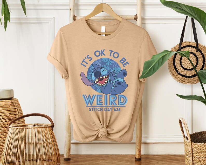 Puede incluir: Camiseta color beige con un dise&ntilde;o del personaje Stitch de Lilo & Stitch. El dise&ntilde;o incluye el texto "IT'S OK TO BE WEIRD" y "STITCH DAY 626". El personaje de Stitch es azul y sonr&iacute;e.