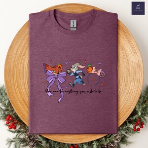 May include: A maroon t-shirt featuring cartoon characters from Zootopia, including Nick Wilde and Judy Hopps, with a purple bow and a carrot headband. The text "You can be anything you wish to be" is printed below the characters.