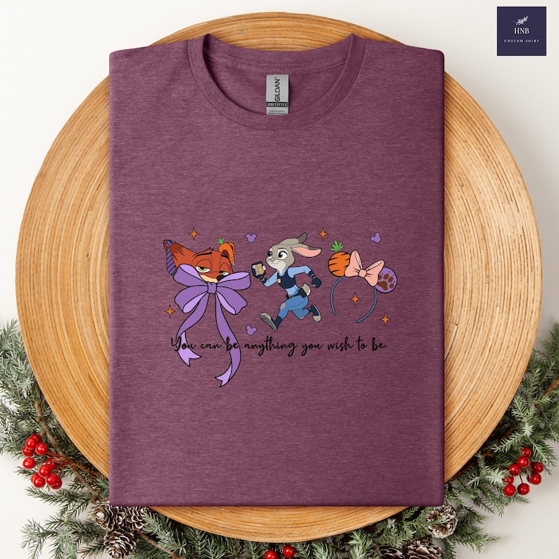 May include: A maroon t-shirt featuring cartoon characters from Zootopia, including Nick Wilde and Judy Hopps, with a purple bow and a carrot headband. The text "You can be anything you wish to be" is printed below the characters.