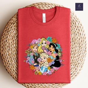 May include: Red t-shirt featuring a floral design with cartoon princesses. The design includes Anna, Elsa, Rapunzel, and Jasmine, surrounded by colorful flowers. The shirt has a crew neck and a tag that says "Canvas".