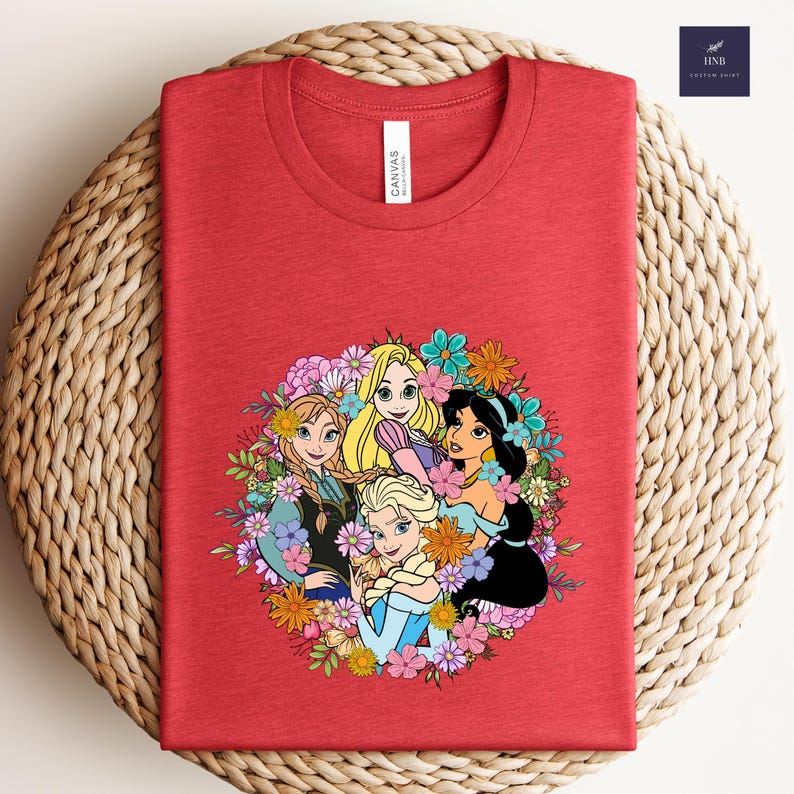 May include: Red t-shirt featuring a floral design with cartoon princesses. The design includes Anna, Elsa, Rapunzel, and Jasmine, surrounded by colorful flowers. The shirt has a crew neck and a tag that says "Canvas".