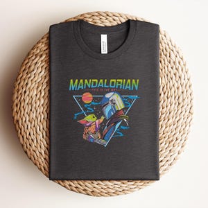 May include: Dark grey t-shirt with a vibrant graphic of The Mandalorian and Grogu, with the text "MANDALORIAN" and "THIS IS THE WAY". The design features a helmeted figure and a small green character, set against a space-themed background.