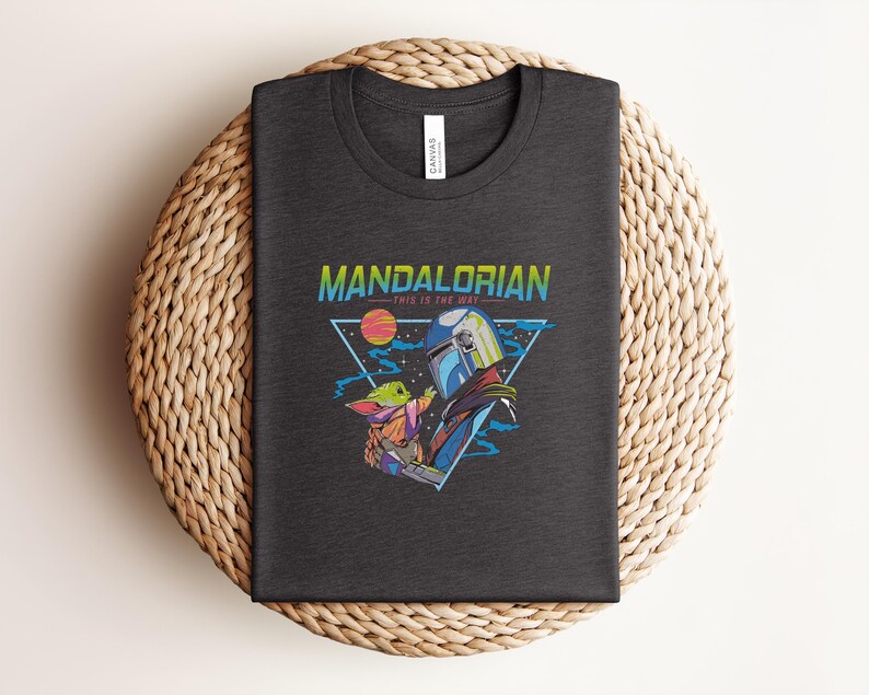 May include: Dark grey t-shirt with a vibrant graphic of The Mandalorian and Grogu, with the text "MANDALORIAN" and "THIS IS THE WAY". The design features a helmeted figure and a small green character, set against a space-themed background.
