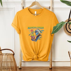 May include: Mustard yellow t-shirt with a graphic of the Mandalorian and Grogu. The design includes the text "Mandalorian - This is the Way" in green and blue. The shirt is hanging on a wooden hanger.