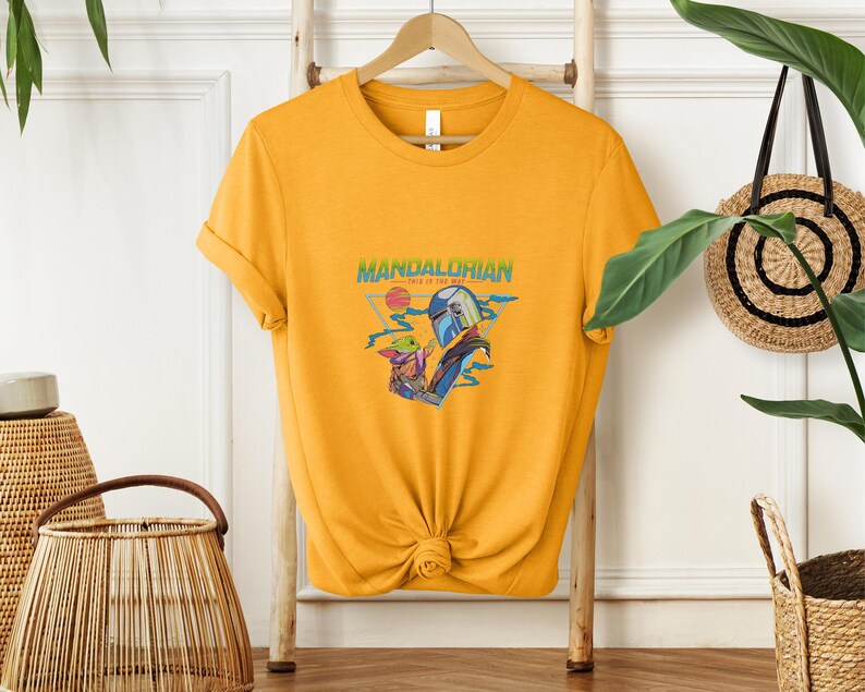 May include: Mustard yellow t-shirt with a graphic of the Mandalorian and Grogu. The design includes the text "Mandalorian - This is the Way" in green and blue. The shirt is hanging on a wooden hanger.